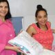 Mulher em maca hospitalar com recém-nascida ao lado de mulher de com jaleco rosa entregando um pacote.