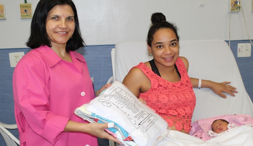 Mulher em maca hospitalar com recém-nascida ao lado de mulher de com jaleco rosa entregando um pacote.