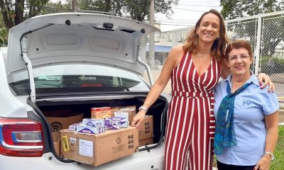 Carro com o porta-malas aberto, aparentemente, várias caixas de produtos de higiene; ao lado direito, uma senhora de cabelo curto com uniforme do hospital são vicente abraçada com uma mulher alta de macacão branco e vermelho listrado