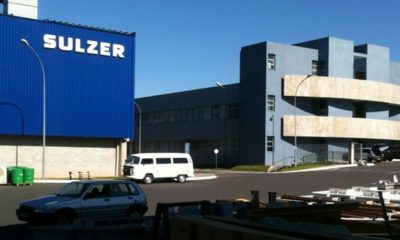Prédio azul da empresa Sulzer, com veículos branco e cinza estacionados na frente da empresa