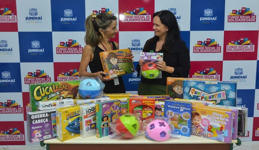 Mulher loira, Maria Aparecida Firigatte de Ares, segurando jogos de mesa ao lado de mulher de cabelos escuros, Vanessa Machado também segurando brinquedo em mãos