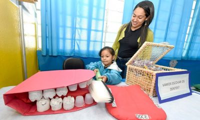 Menina escovando dentes de uma boca gigante de gesso, ao lado de uma mulher de cabelos escuros e com jaqueta verde