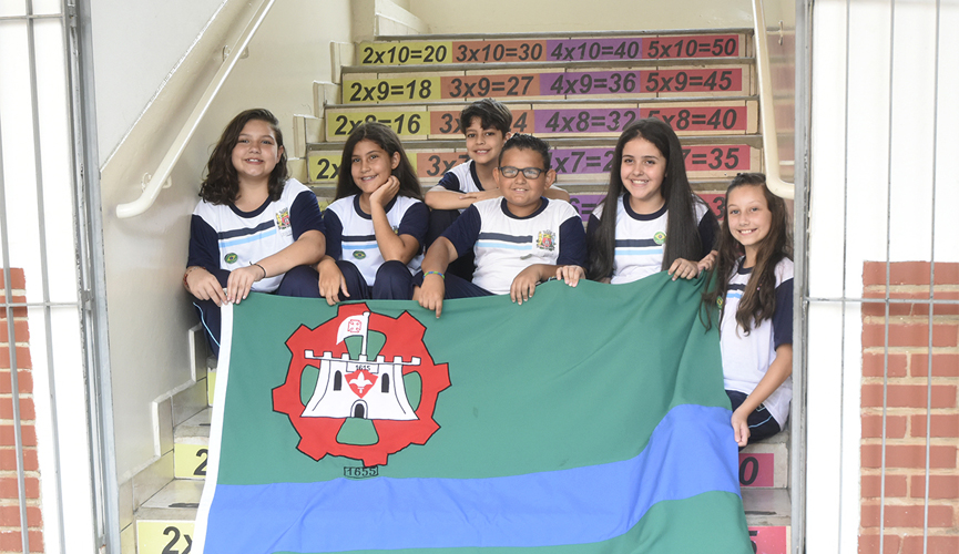 Seis crianças, sendo quatro meninas e dois meninos, sentados em escada com números da tabuada escritos e segurando a bandeira de Jundiaí