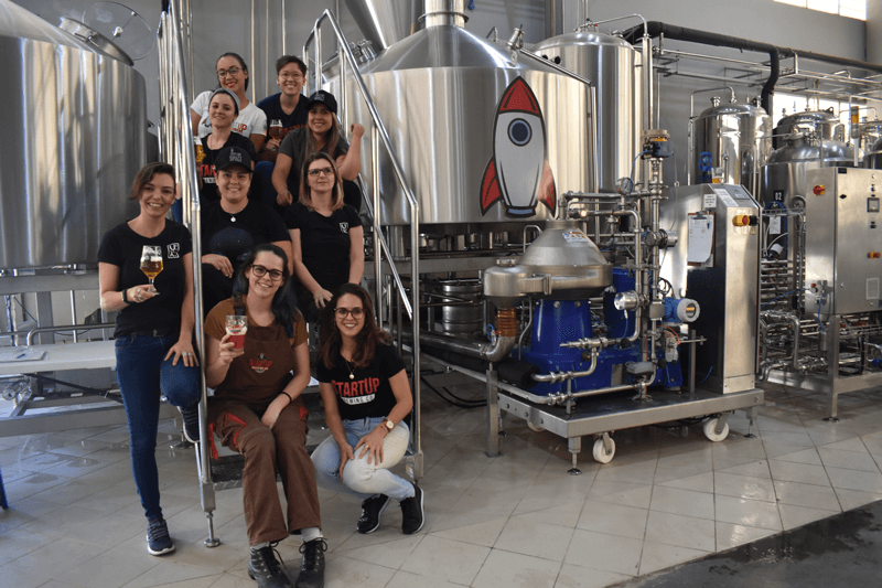 Mulheres colaboradoras da cervejaria StartUp Brewing, sentadas em meio ao maquinário da cervejaria
