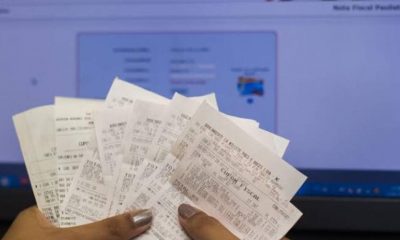 Mão segurando notas fiscais com tela de computador com site da nota fiscal paulista aberto