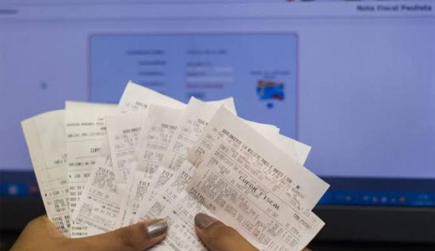 Mão segurando notas fiscais com tela de computador com site da nota fiscal paulista aberto