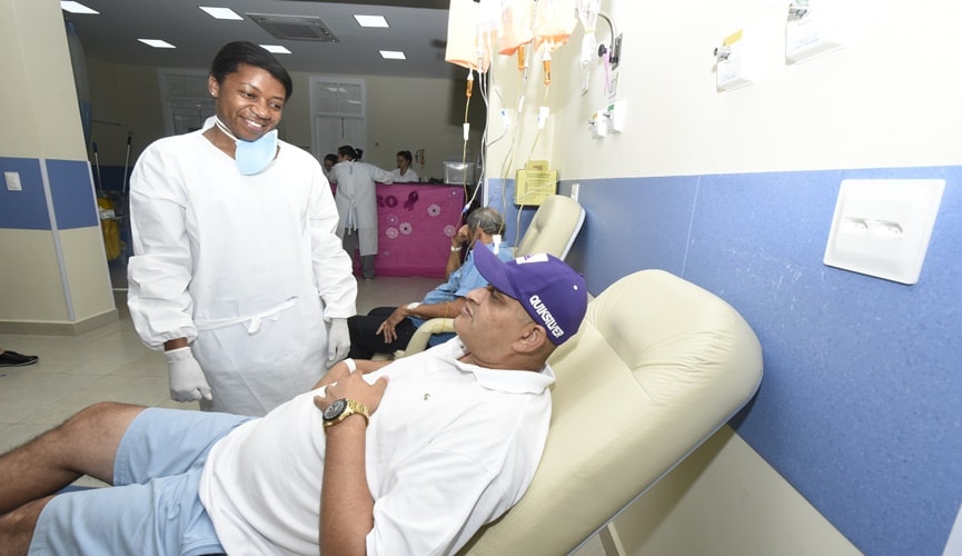 Ala de quimioterapia, com enfermeira negra com trajes brancos, de pé à frente de paciente pardo, de camisa polo branca, bermuda jeans e boné azul,que recebe medicamento,