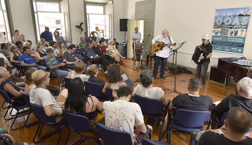 Músicos idosos fazendo show em uma sala com jovens, adultos e idosos.