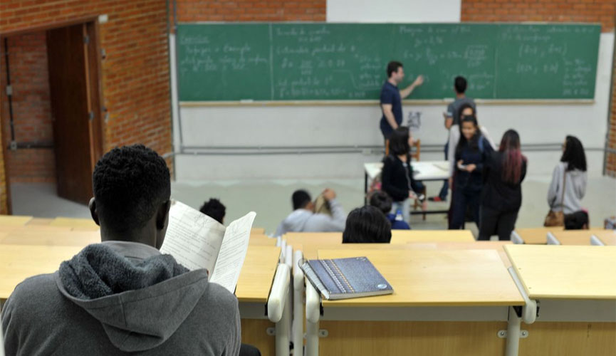 estudante negro em sala de aula, sentado em mesa. Ao fundo, outros estudantes aparecem desfocado ao lado do professor e da lousa