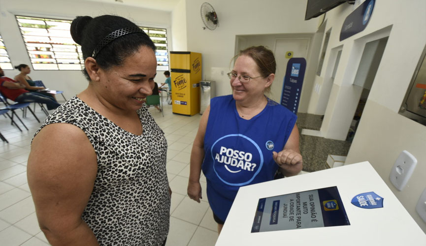 Mulher observa paineld e votação em unidade de saúde, enquanto funcionária aparece sorridente para ajudá-la
