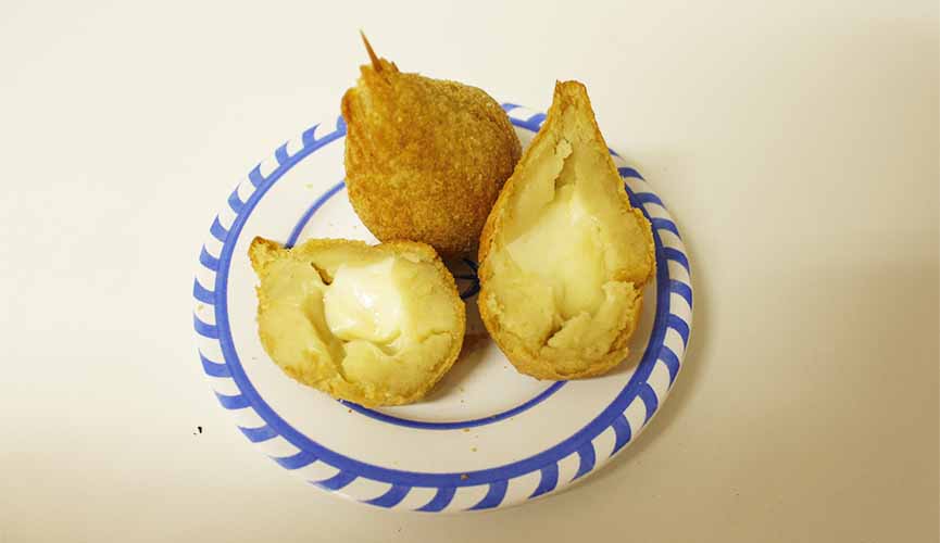 Duas coxinhas de queijo dentro de um prato. Uma delas dividida ao meio, com as duas partes deitadas e a outra, inteira, com um palito fincado na ponta.