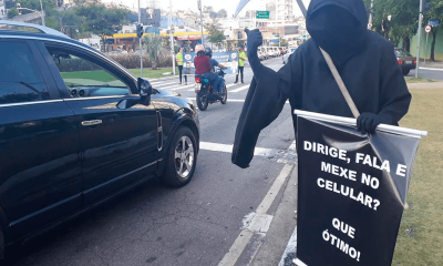 Ator vestido de morte, com capa preta e foice, exibe um cartaz na Av. 9 de Julho: “Dirige, fala e mexe no celular? Que ótimo!”