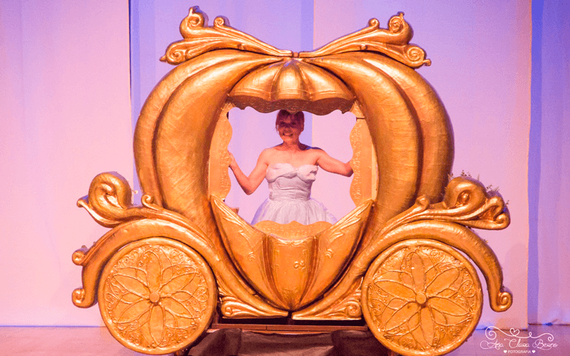 Foto de trecho do espetáculo em que Cinderela está dentro de uma carruagem que se assemelha a uma grande abóbora.
