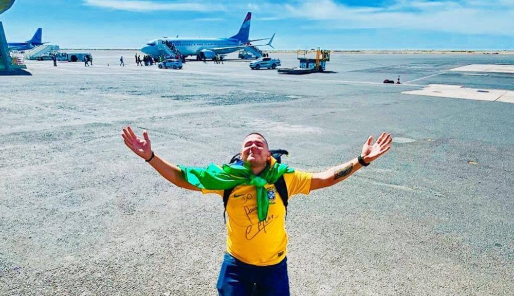 Foto de Anderson ajoelhado em um pátio de aviões vestindo camisa brasileira, com bandeiras enrolada no pescoço. Ele ergue as mãos para o céu como forma de agradecimento