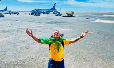 Foto de Anderson ajoelhado em um pátio de aviões vestindo camisa brasileira, com bandeiras enrolada no pescoço. Ele ergue as mãos para o céu como forma de agradecimento