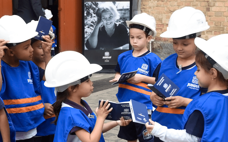 Crianças da rede municipal em roda, com capacete de proteção, segurando seus passaportes culturais