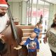Criança com câncer tocando em instrumento de músico com gorro de Papai Noel, ao lado da mãe