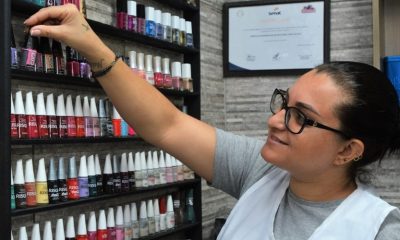 Mulher de óculos, com jaleco branco, escolhendo esmalte em prateleira fixada em parede com textura