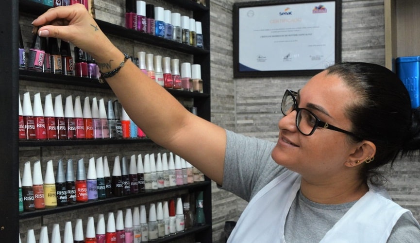 Mulher de óculos, com jaleco branco, escolhendo esmalte em prateleira fixada em parede com textura