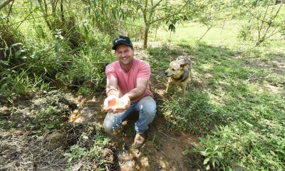 produtor rural mostra água limpa de nascente após trabalho de preservação. ele está em meio a mata, segurando um pouco da água. há um cachorro atrás dele