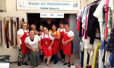 voluntárias do bazar do hospital são vicente com aventais vermelhos debaixo da entrada... logo acima, na parede, está escrito "bazar do voluntariado visita vicente, sejam bem vindos". no canto da imagem é possível ver roupas penduradas em cabides