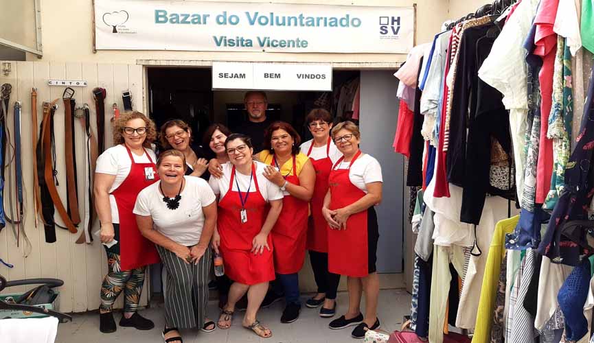 voluntárias do bazar do hospital são vicente com aventais vermelhos debaixo da entrada... logo acima, na parede, está escrito "bazar do voluntariado visita vicente, sejam bem vindos". no canto da imagem é possível ver roupas penduradas em cabides