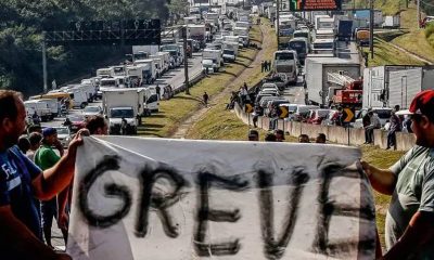 dois caminhoneiros seguram, cada um em uma ponta, uma faixa escrito "GREVE". Ao fundo, duas mãos de rodovia estão completamente tomadas por caminhões estacionados, durante greve de 2018