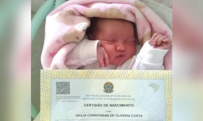 bebê deitada em cama dormindo com certidão de nascimento a sua frente, com o nome "giulia corinthiana de oliveira costa