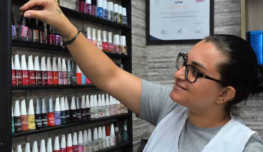 manicure mexendo em painel com uma série de esmaltes fixado na parede. ao fundo, um quadro com seu diploma