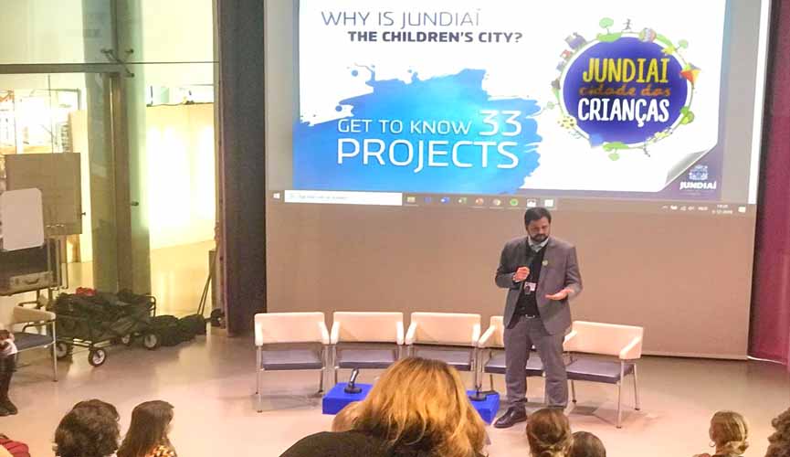 prefeito de jundiaí com microfone em auditório com slide atrás onde diz, em inglês: "´porque jundiaí é a cidade das crianças?"