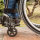 imagem da close em roda de cadeira de rodas e em perna de deficiente. foto é lateral e mostra pedaço da perna e do sapato, assim como pedaço da roda
