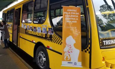 Ônibus urbano de Jundiaí em campanha contra violência a mulher, com banner "Jundiaí pela fim da violência contra a mulher, denuncie 180". Mais atrás, também na lateral da parte externa do ônibus, imagens de conscientização formal um varal de fotos