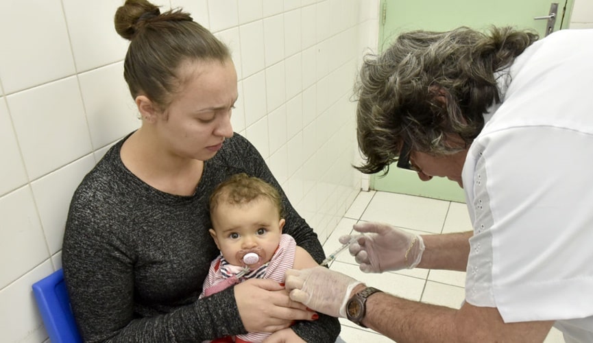 Foto de criança sendo vacina no colo da mãe por enfermeira idosa