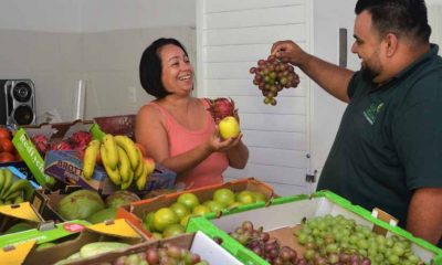 homem e mulher seguram frutas atrás de mesa cheia de caixotes com frutas e legumes