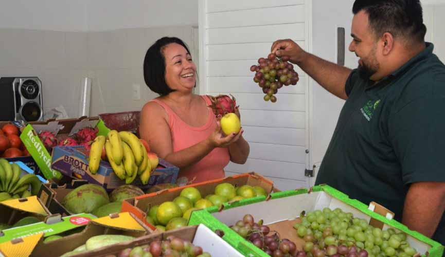 homem e mulher seguram frutas atrás de mesa cheia de caixotes com frutas e legumes