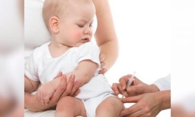 Segurado pela mãe, bebê observa enquanto mãos de enfermeira aplicam vacina em sua perna esquerda