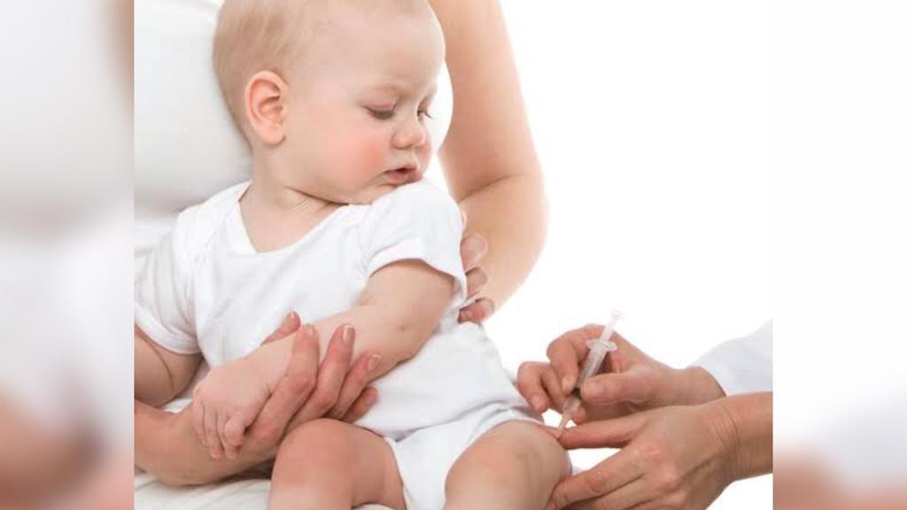 Segurado pela mãe, bebê observa enquanto mãos de enfermeira aplicam vacina em sua perna esquerda