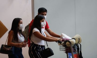 Duas mulheres morenas de cabelos longos pretos, com homem moreno de cabelo curto, saindo de aeroporto com máscaras