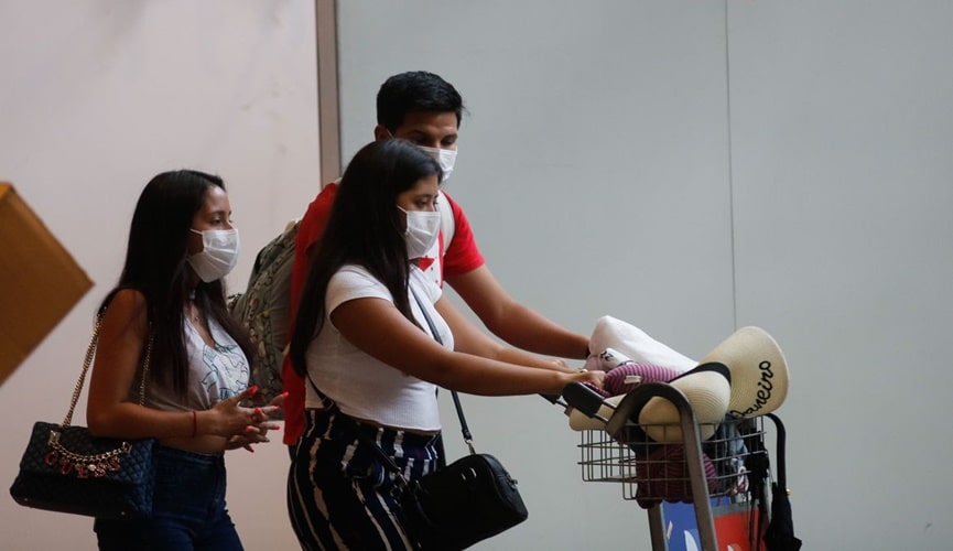 Duas mulheres morenas de cabelos longos pretos, com homem moreno de cabelo curto, saindo de aeroporto com máscaras