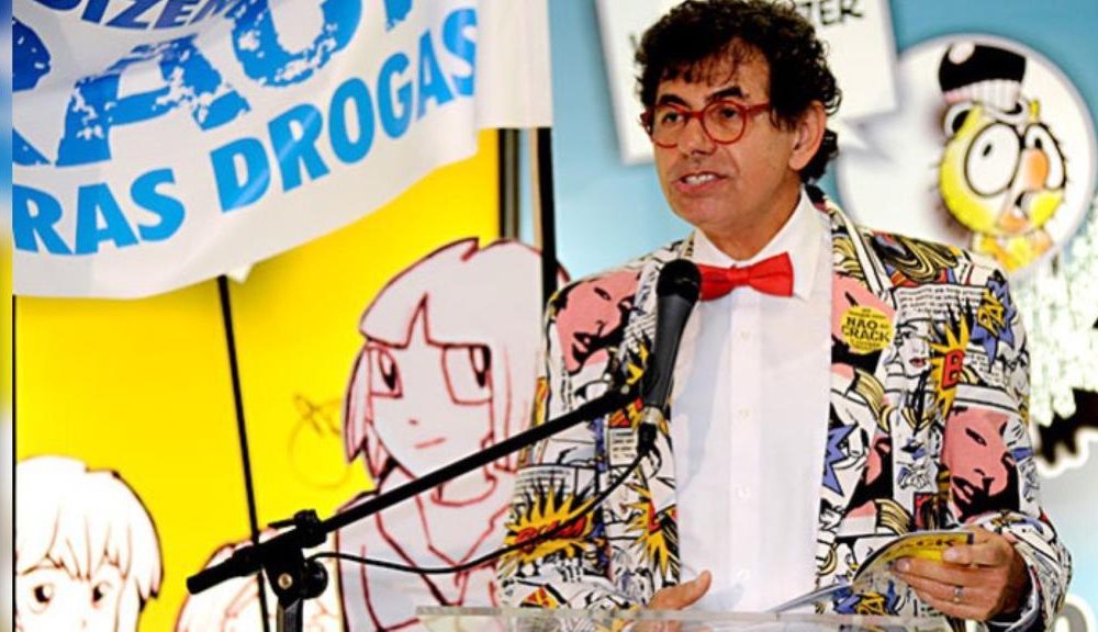 De óculos, com gravata borboleta e blazer de história em quadrinhos, Daniel Azulay está em pé falando ao microfone