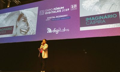 Paola Faria palestrando no palco da Expo Fórum Digital 2019; ela está com blazer bege, calça preta, camiseta rosa e tênis branco, segurando microfone na frente de painel roxo.