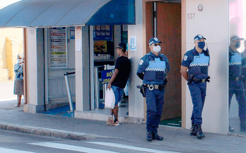 Guardas municipais de máscara andam por rua, próximo a lotérica