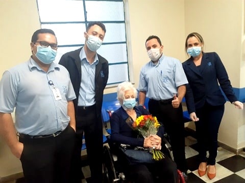 Foto de idosa com buquê de flores e equipe médica ao lado