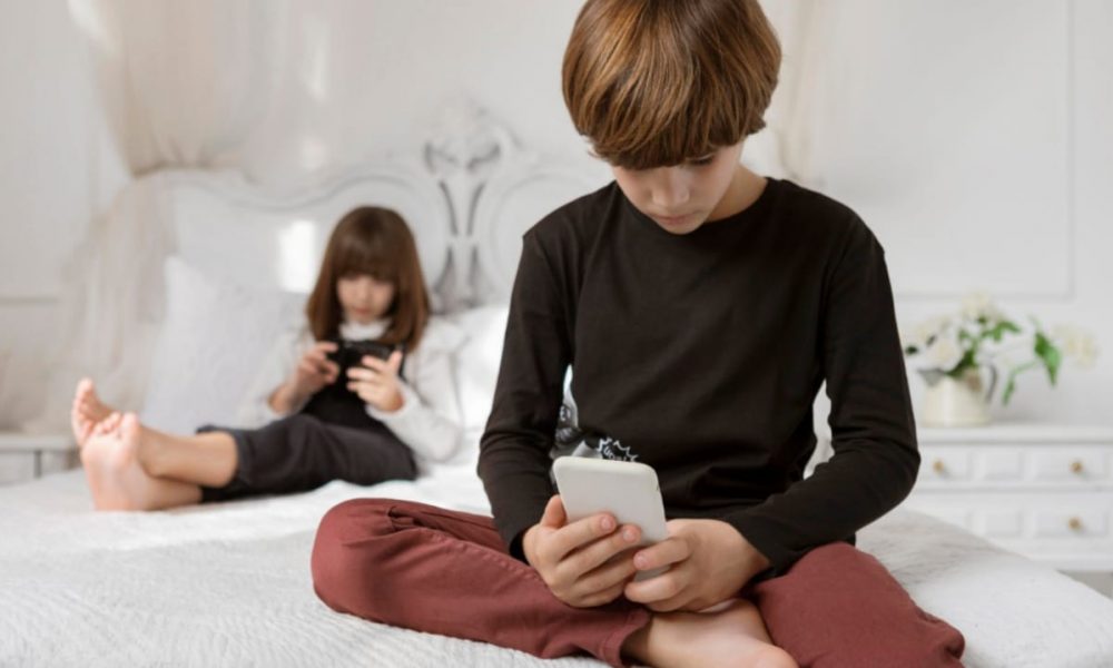 Duas crianças sentadas na cama com celulares na mão