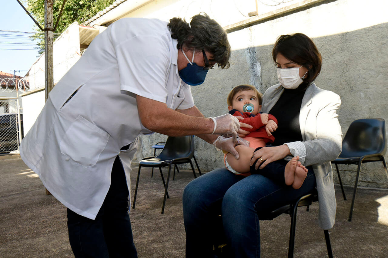 auxiliar de enfermagem aplicando vacina em uma criança sendo segurada pela mãe