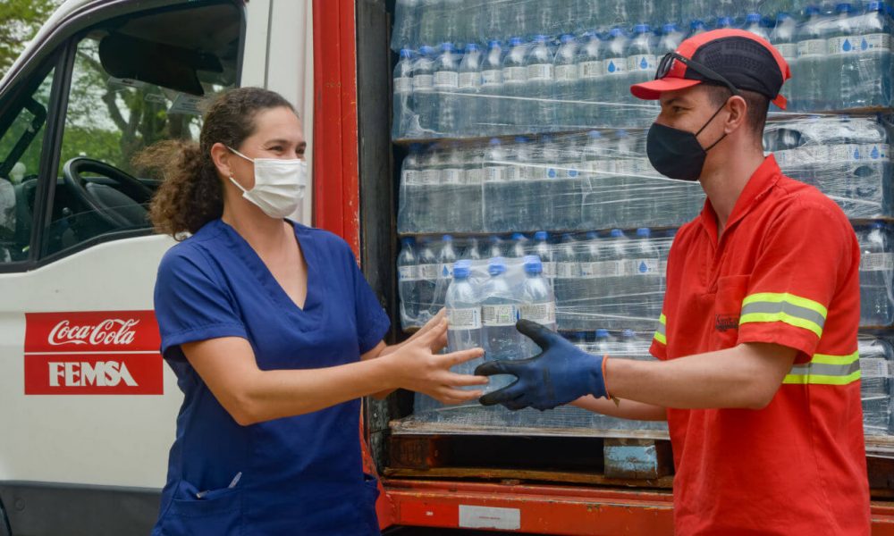 um homem com roupas da coca cola entregando um fardo de garrafas de água para uma mulher, com um caminhão atrás