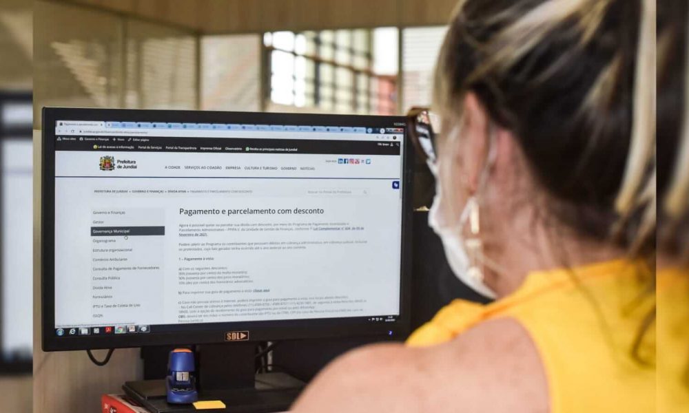Uma mulher de blusa amarela sentada na frente do computador que está aberto no site da prefeitura de Jundiaí