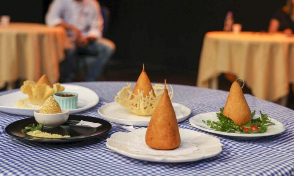 A imagem mostra uma mesa coberta com uma toalha de mesa xadrez azul e branca, sobre a qual estão dispostas várias coxinhas de queijo apresentadas em pratos decorativos. As coxinhas têm formato cônico e algumas estão acompanhadas de guarnições como folhas verdes e molhos. Ao fundo, há outras mesas com toalhas de cor clara e pessoas desfocadas, sugerindo um ambiente de restaurante ou evento gastronômico.