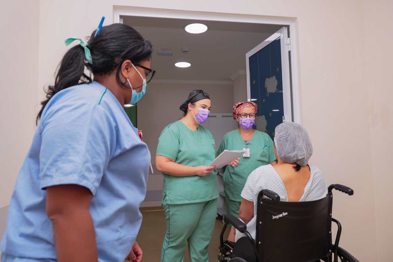 Paciente sentada em uma cadeira de rodas, ao lado de 3 enfermeiras, sendo levada para o centro cirúrgico
