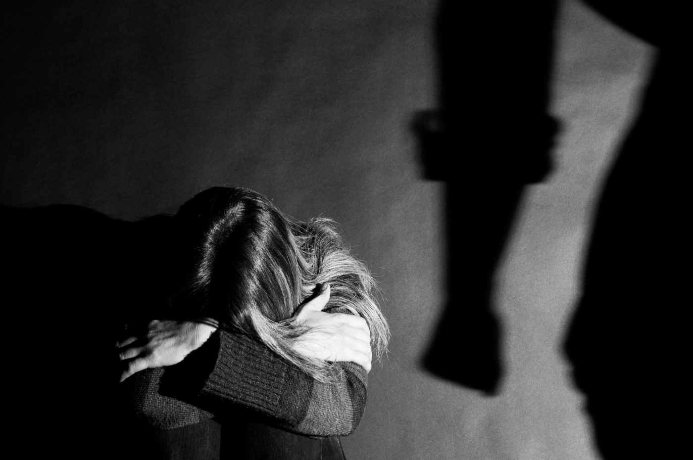 violência contra mulher, uma mulher sentada no chão com a cabeça abaixada e a sombra de um homem com o punho fechado ameaçando
